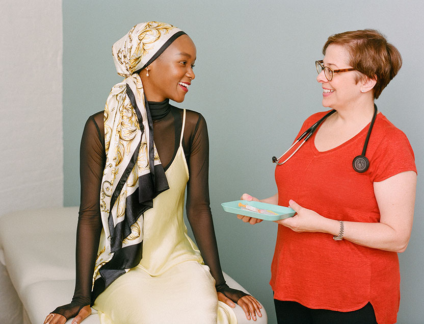 Two women in a doctor's office, looking at each other and smiling. The doctor holds a small tray with a syringe on it.