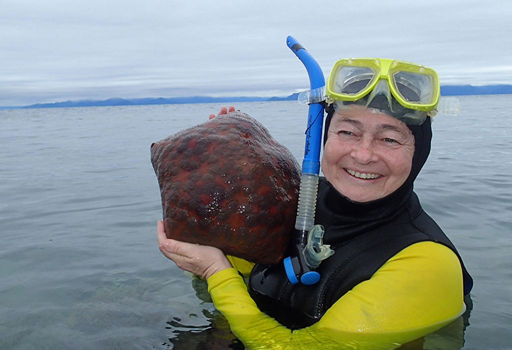 A person (Maria Byrne) standing in calm water in snorkelling gear, smiling and holding a sea star