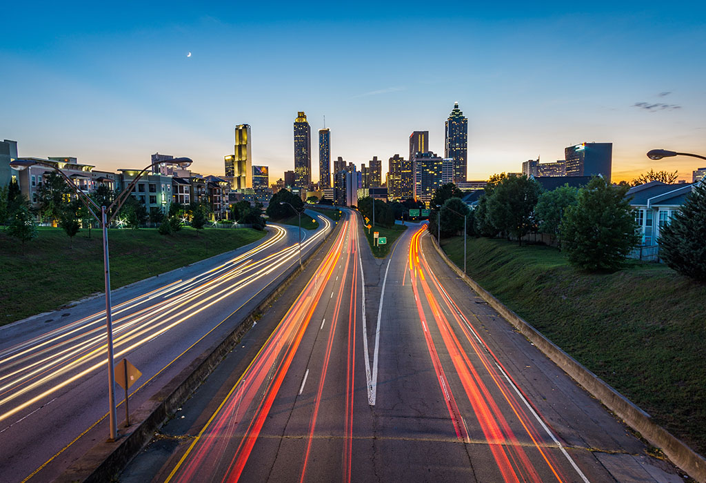 A city skyline in the evening dusk, showing roads leading in and out, with blurry trails of car lights.