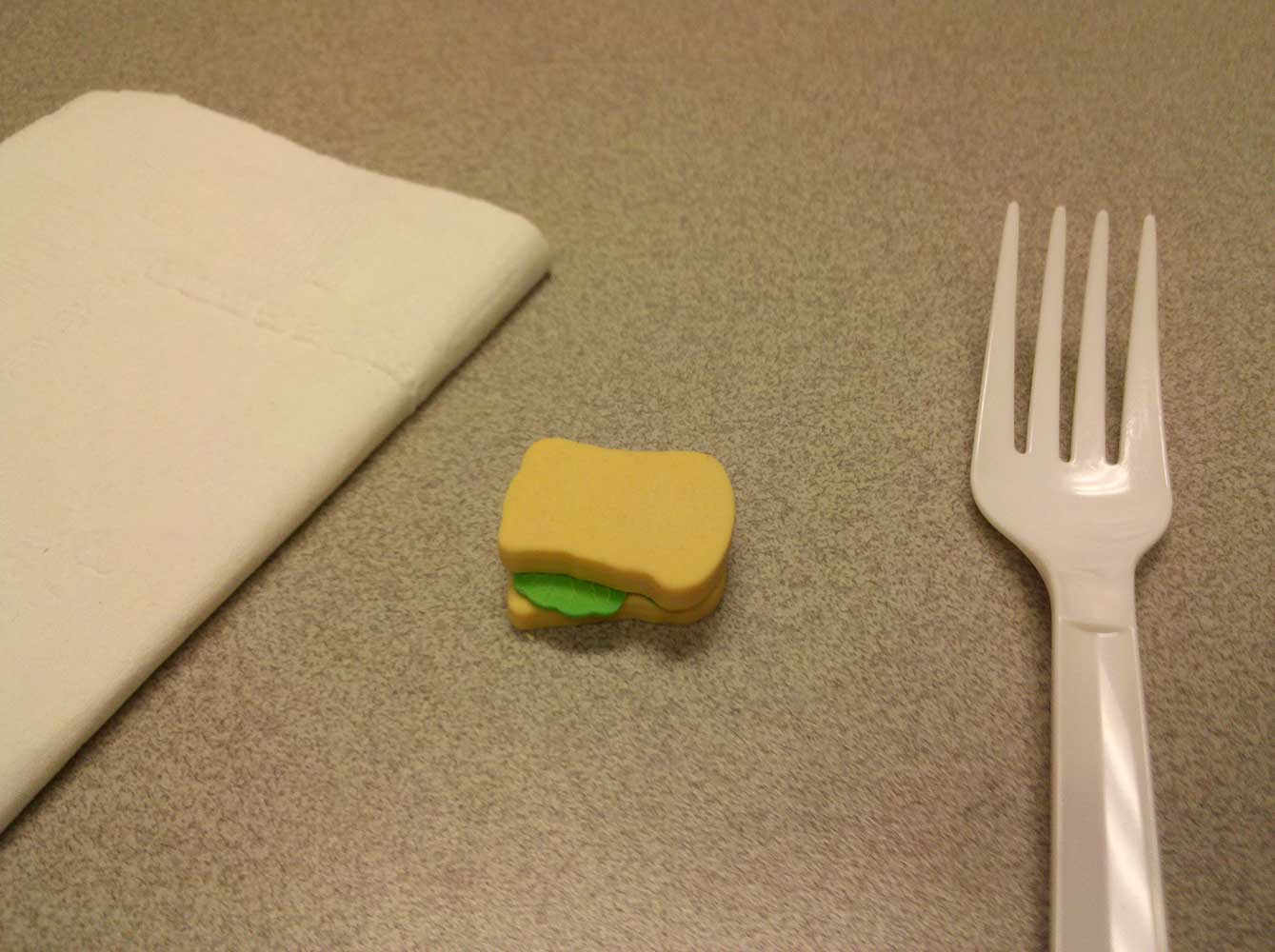 A tiny sandwich next to a fork and napkin which comparatively, look giant.