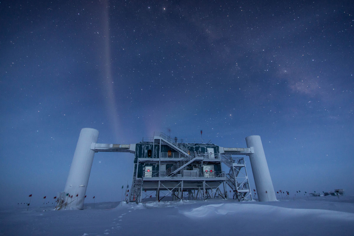 The IceCube neutrino observatory, a large building with detached towers on each side, stands in a snowy plain at night.
