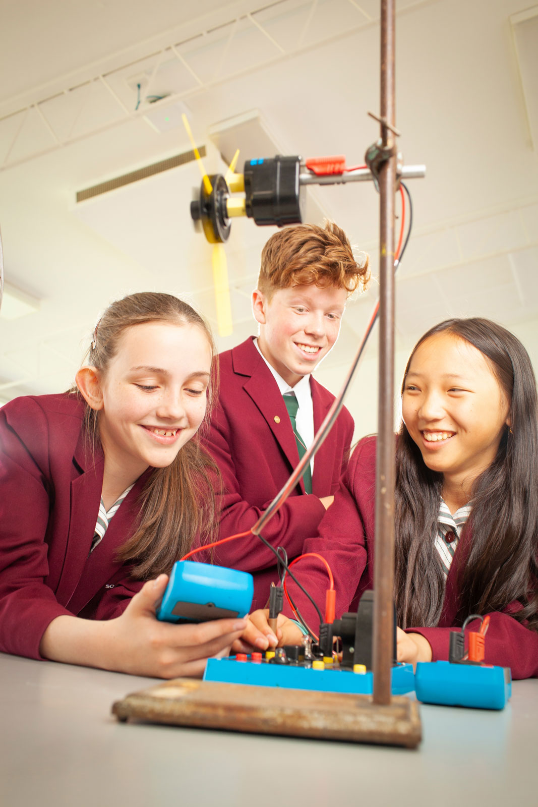 Three students in school uniforms gather around a small electronic meter.