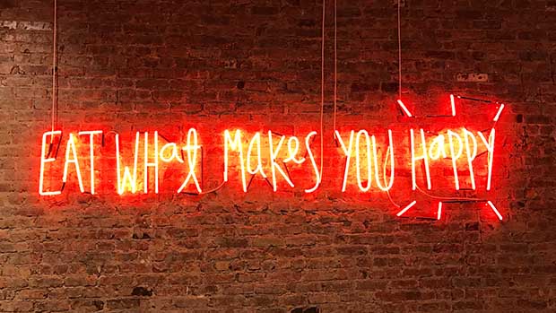 A photo of a neon sign on a brick wall that reads eat what makes you happy.