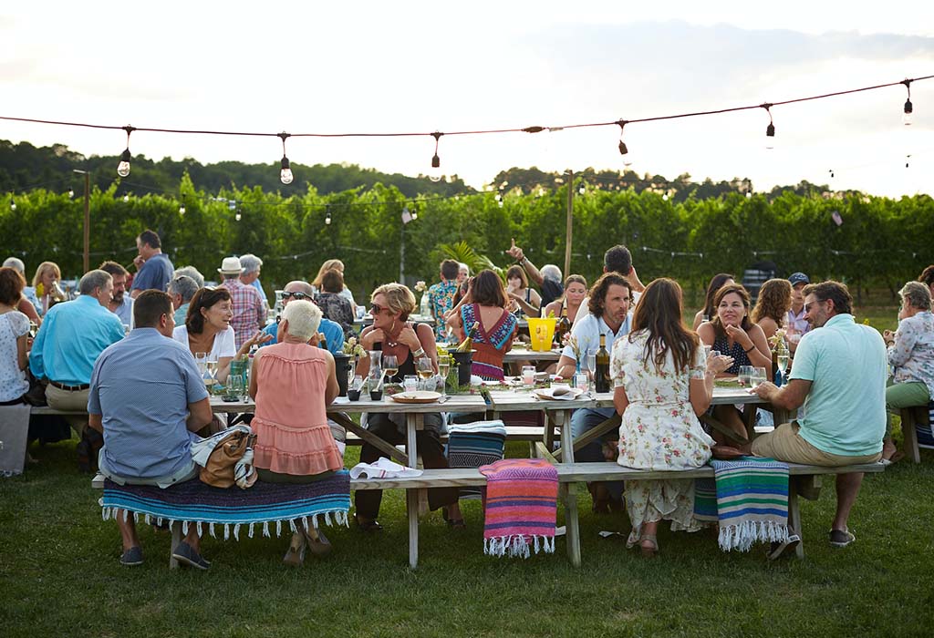 Some long dining tables and chairs, outdoors on a lawn, with many people enjoying a meal together.