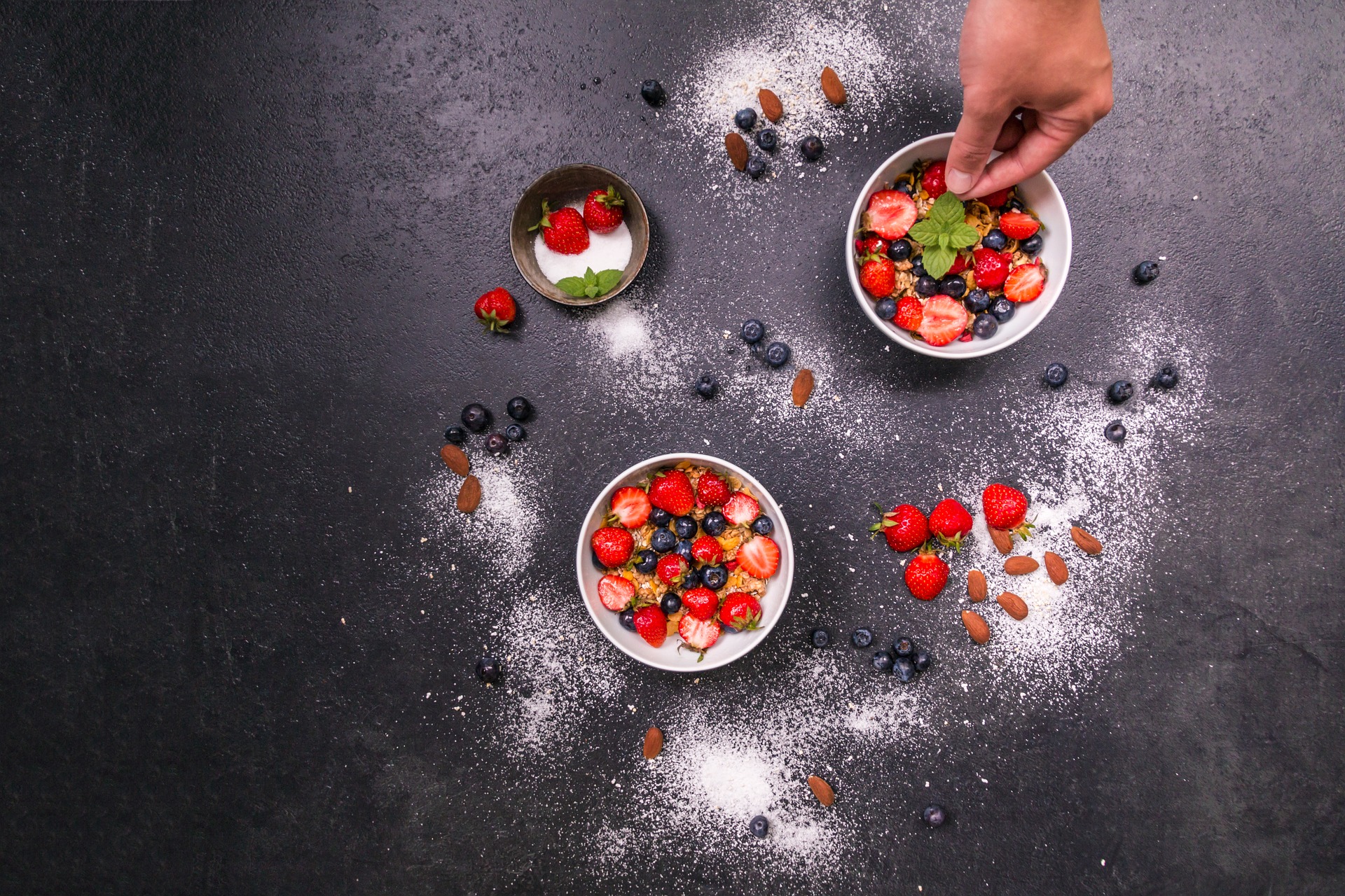 Hands reaching towards some small bowls of berries and cereal on a dark surface