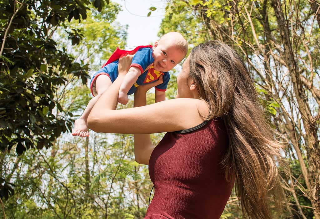 A mother holding up her baby, who wears a Superman costume