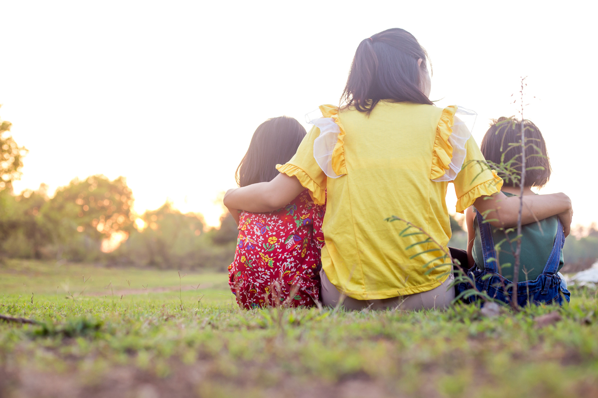 A mother hugs two children, they are sitting in a park looking towards the sunset, back to camera.