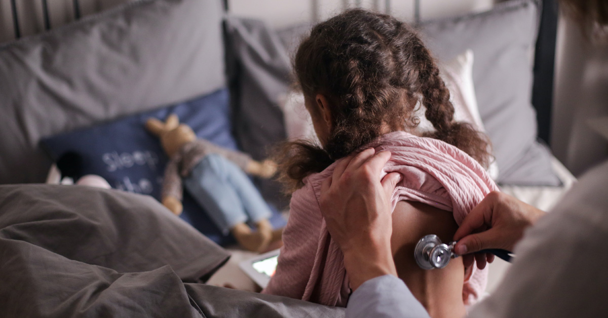 A child with her back to the camera is being examined by a person with a stethoscope.