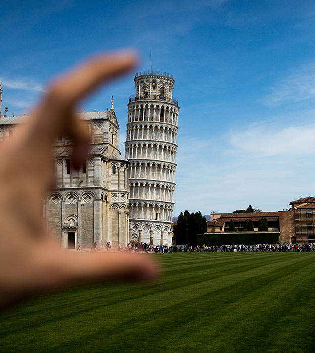 The Leaning Tower of Pisa on a clear sunny day, with a person’s hand in the foreground of the image, appearing to hold the tower between two fingers.