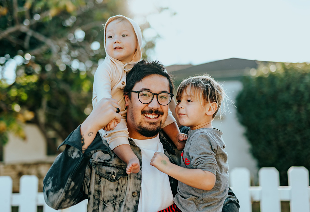 A parent with his two children. One sits on his shoulders, the other is in his arms.