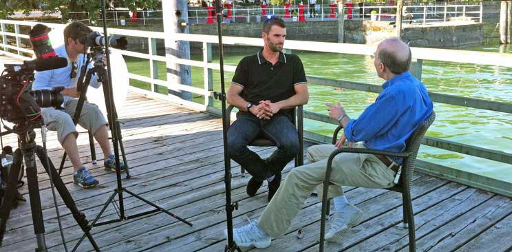 Two men and a photographer sitting on a chairs on a wharf, with photo and video equipment in the foreground