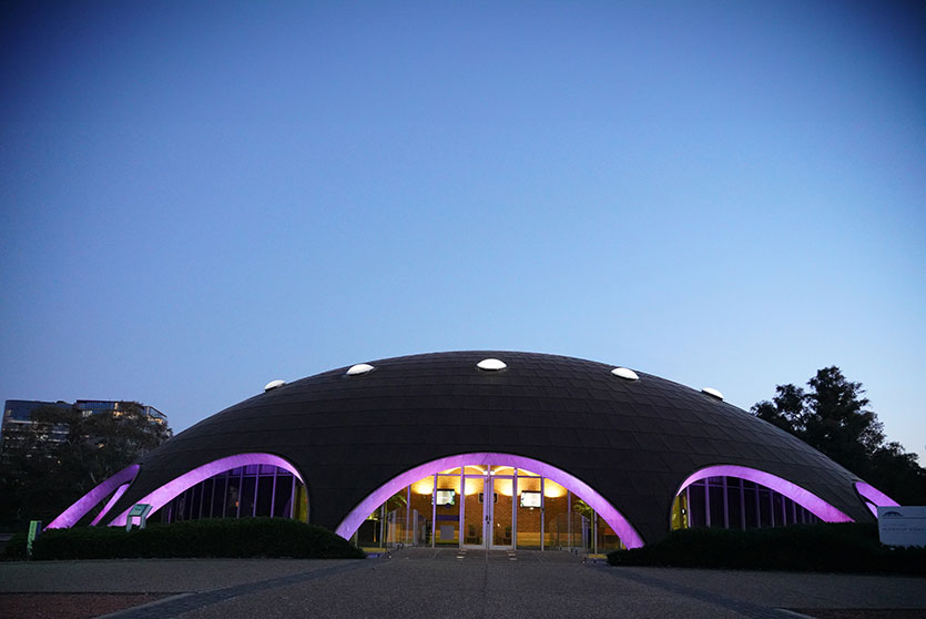 The Shine Dome building lit up with purple under its arches beneath a dark blue sky at dusk