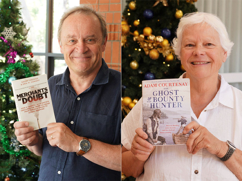 A collage of two images: on the left, a man holds up a book titled Merchants of Doubt. He is smiling at the camera and standing in front of a Christmas tree. On the right, a woman holds up a book titled The Ghost and the Bounty Hunter. She is also smiling and standing in front of a Christmas tree. 