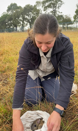 Woman kneeling in a grassy field showing a small mammal curled up in a white cloth bag