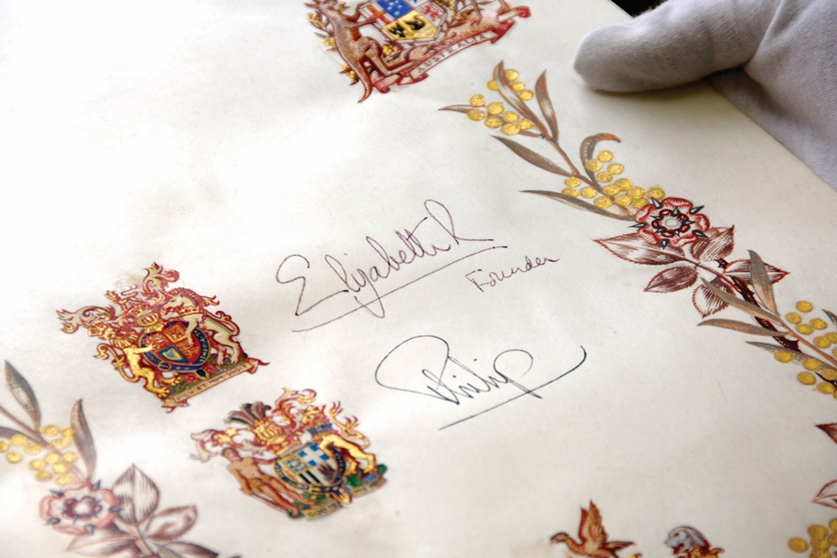 A gloved hand holds a colored embossed page from a book which bears the signatures of 'Elizabeth R (Founder)' and 'Philip'