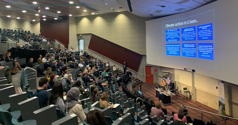 Large modern lecture theatre about half full of people, with a presenter in front of a large projected slide that says 'Climate action in 6 boxes'