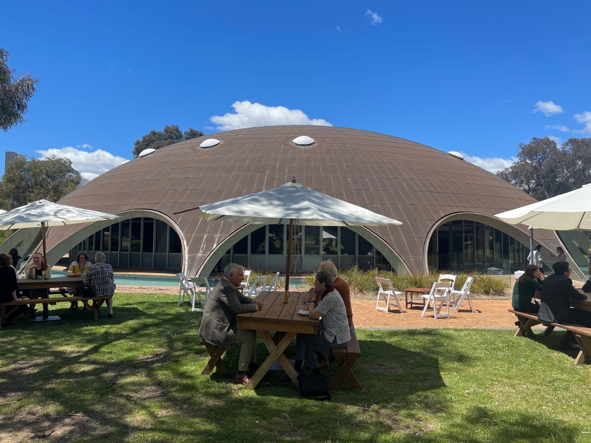 People sit outside at picnic tables in front of a large copper domed building 