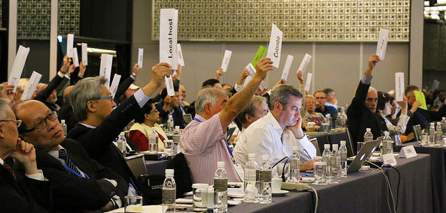 People seated in rows, with their hand up holding their country's name