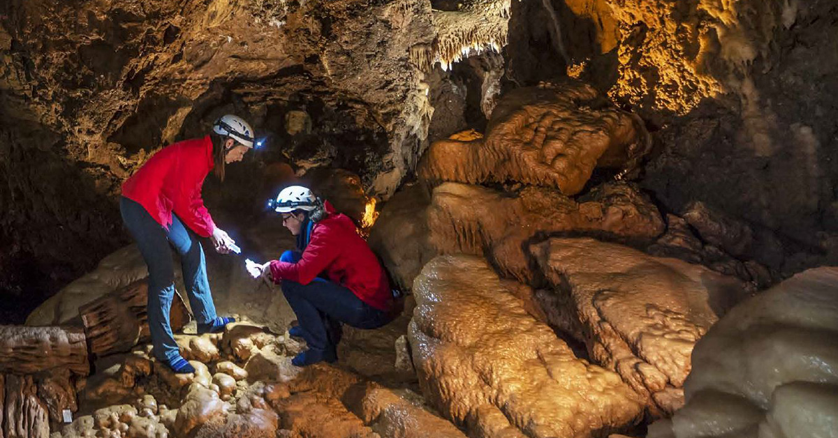 Two women in a limestone cave wearing helmets with head torches and holding small bottles