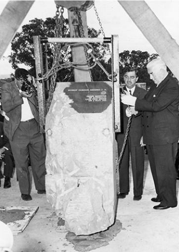 Large stone being lowered into place, with several men in suits nearby