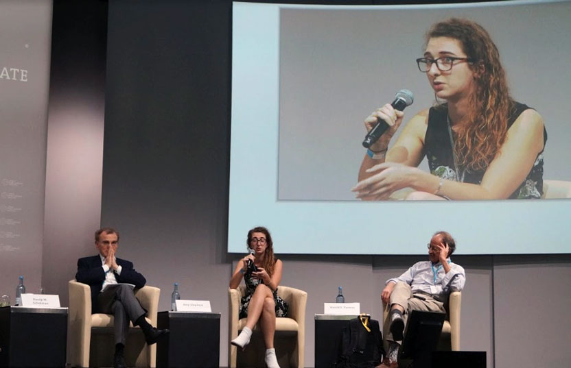 Woman speaking into a microphone, seated on stage with two men, and a large screen behind them with a close-up of her speaking