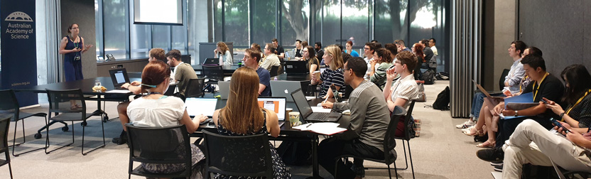 Speaker in front of large group of young adults in a conference room who are seated at tables using laptops