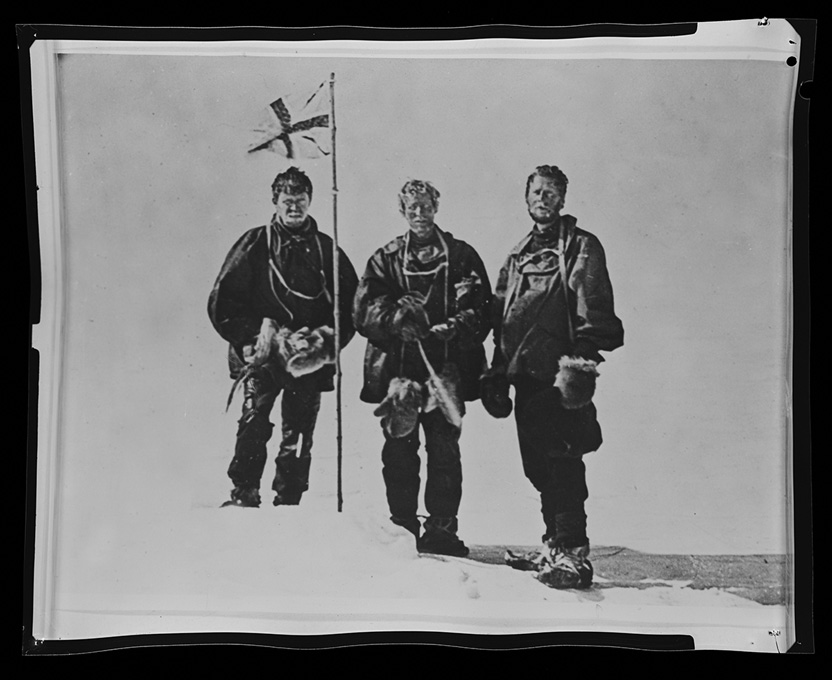 Black and white photo of three men in clothing to protect against the cold standing on an icy peak with a flag pole stuck in the snow.
