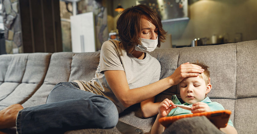 Woman wearing a COVID mask on a lounge, looking concerned and feeling the forehead of a toddler who is holding and looking at a thermometer