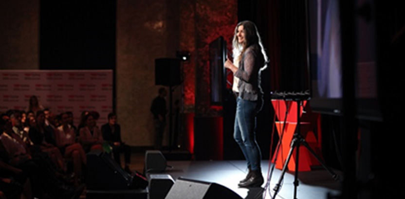 Smiling woman speaking on a stage in front of an audience with a red X in the background