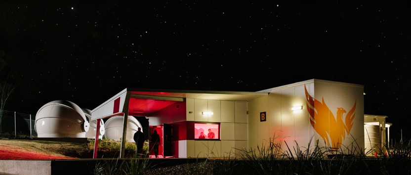 Single storey modern building lit at night, in front of several rounded structures that hold telescopes
