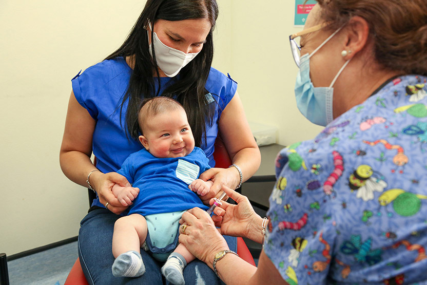 A woman is holding a smiling baby in a doctor's office. The woman doctor is giving the baby an injection.