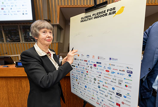 Professor Lidia Morawska holds a pe ready to sign a posterboard titled 'Global pledge for healthy indoor air' with a canary symbol and a large number of organisations logos on it