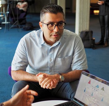 Man wearing glasses sitting at a low round table and in conversation with others out of shot