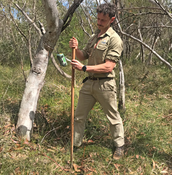 Man in outdoor clothing and boots in a grassy woodland environment holding a long stick with an electronic device on it which is aimed at the ground