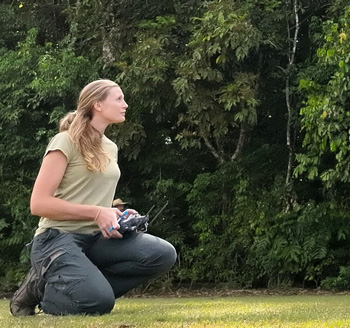 Woman crouched on the ground holding a drone controller and looking up, with a wall of thick green tree vegetation in the background.