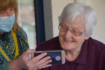 A elderly woman looks at a phone screen held by another woman wearing a mask. Image by Photo by Georg Arthur Pflueger on Unsplash.