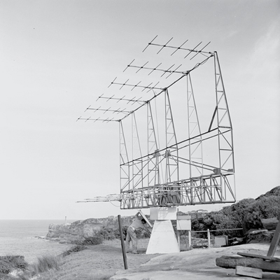 Near the edge of a cliff overlooking the sea is a joined framework of six metal antennae set up on a plinth, with the metal component looking to be around four metres high. A man is reaching into an access hatch on the side of the plinth. Photo is black and white.