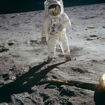 Person completely enclosed in a white spacesuit with a reflecting cover over the face and carrying a white pack on their back, facing the camera and standing on what looks to be an uneven greyish dusty flat landscape with some small rocks and boot-prints in the dust.